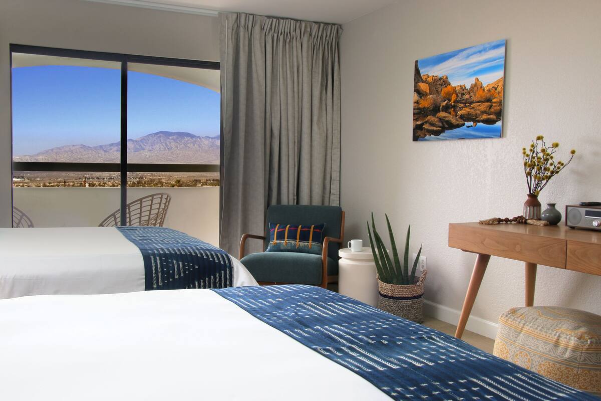 A hotel room with a large window offering a mountain view, a bed with patterned bedding, a chair, a desk with a radio, and a plant.