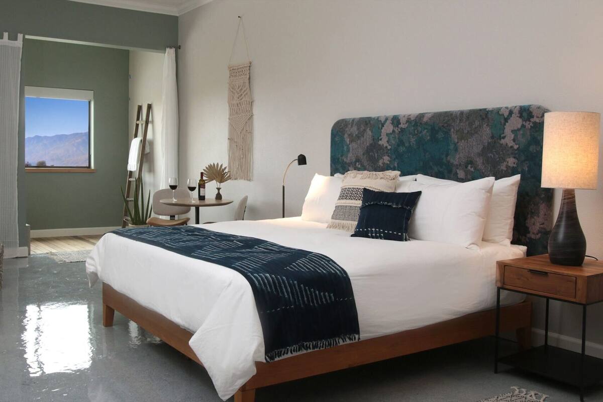 A bedroom with a bed, bedside table, lamp, and a view of the outdoors.