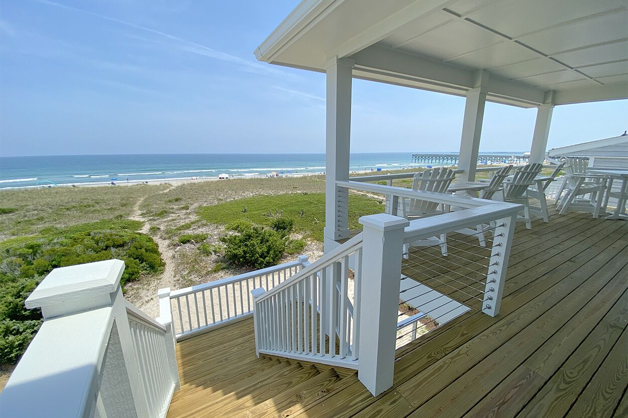 Une terrasse en bois avec des balustrades blanches, donnant sur une plage et offrant une vue dégagée sur l’océan, ainsi qu’une jetée visible au loin.