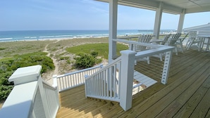 A wooden deck with white railings overlooking a beach with a clear view of the ocean and a pier in the distance.