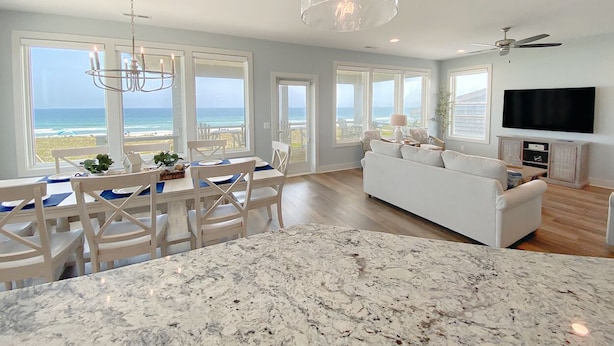A spacious living room with a white sofa, a dining area with a table and chairs, a large window overlooking the ocean, and a flat-screen TV mounted on the wall.