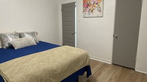 A bedroom with a bed, pillows, a blue bedspread, a beige blanket, a painting, and two doors.
