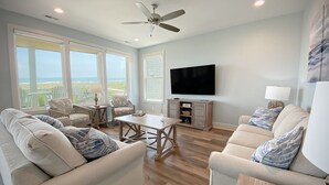Uma sala de estar ampla com uma televisão grande, assentos confortáveis e vista para o oceano.