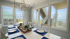 Uma área de jantar com uma mesa branca posta para quatro, jogos americanos azuis e uma peça central com uma cesta. Janelas amplas oferecem vista para o oceano e um edifício com telhado verde.