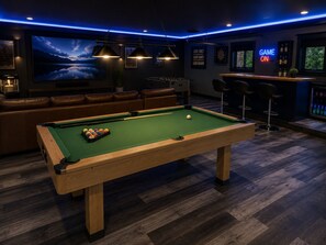 A pool table with balls, a bar area with stools, a large TV screen, and a neon sign.