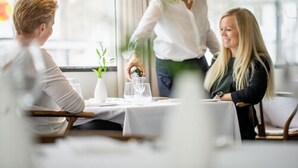 A dining area with a table set for three people, a waiter attending to the guests, and a window providing natural light. - Lindstrøm Hotel (Laerdal)