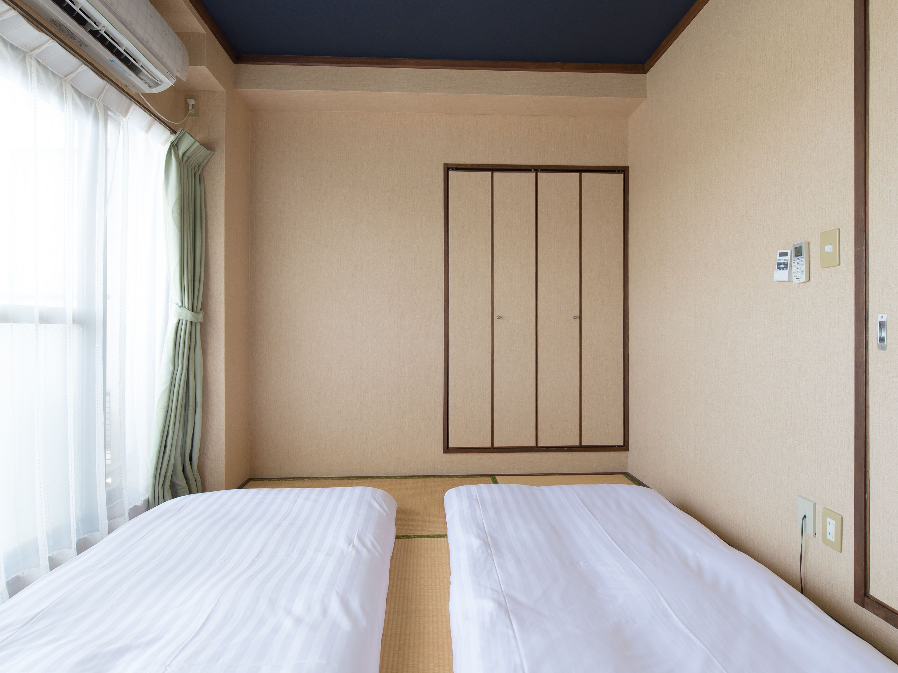 A hotel room with two single beds, a large wardrobe, a window with sheer curtains, and a wall-mounted air conditioning unit.