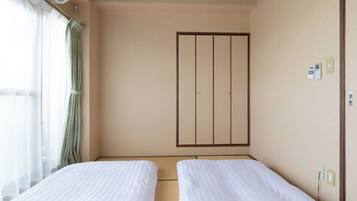 A hotel room with two single beds, a large wardrobe, a window with sheer curtains, and a wall-mounted air conditioning unit.