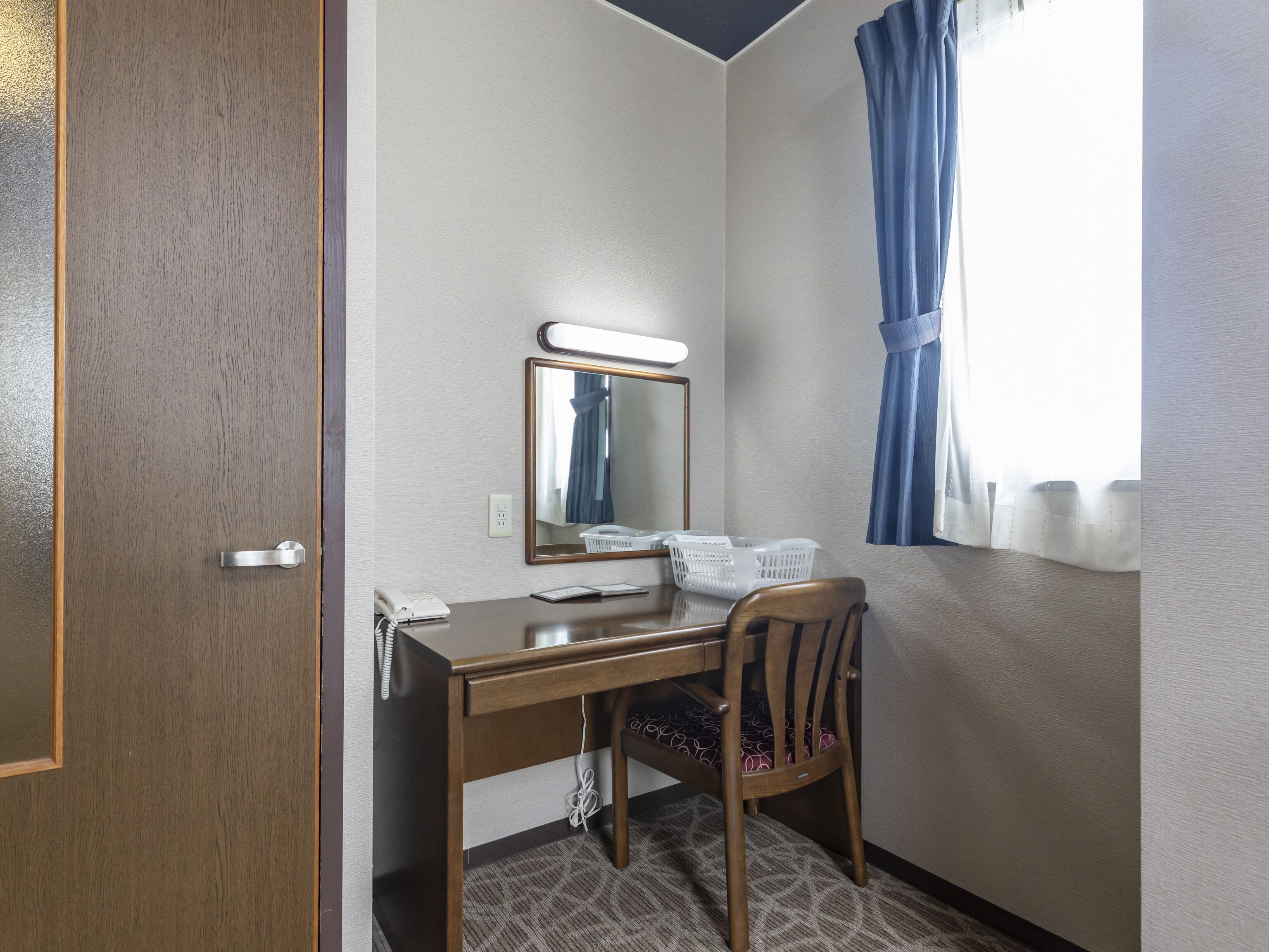 A hotel room with a wooden desk, a mirror, a telephone, and a window with blue curtains.