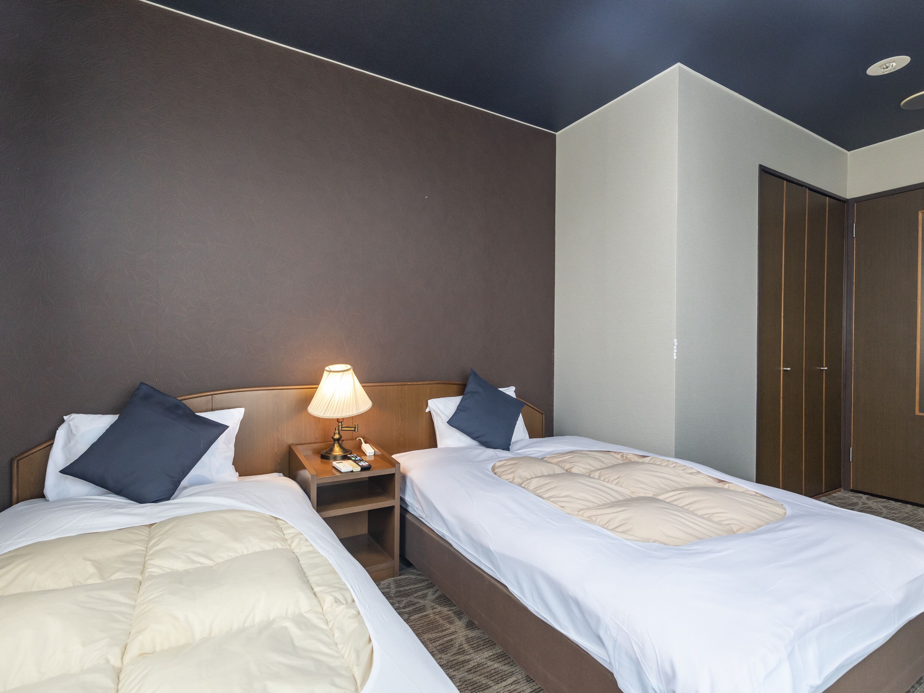 Hotellihuone, jossa on kaksi sänkyä, yöpöytä lampun kanssa ja vaatekaappi.