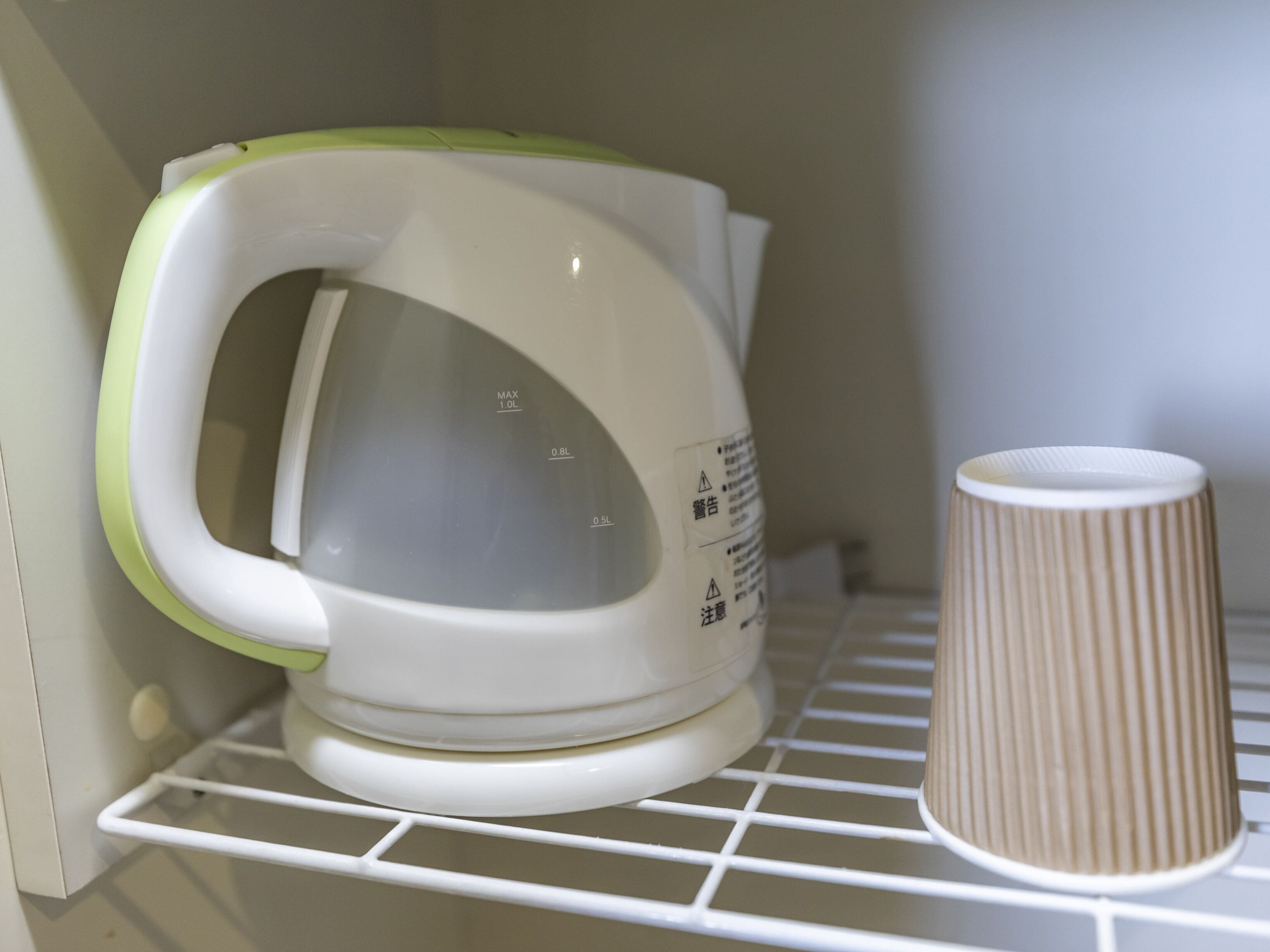 A white kettle with a green accent and a brown paper cup on a white shelf.