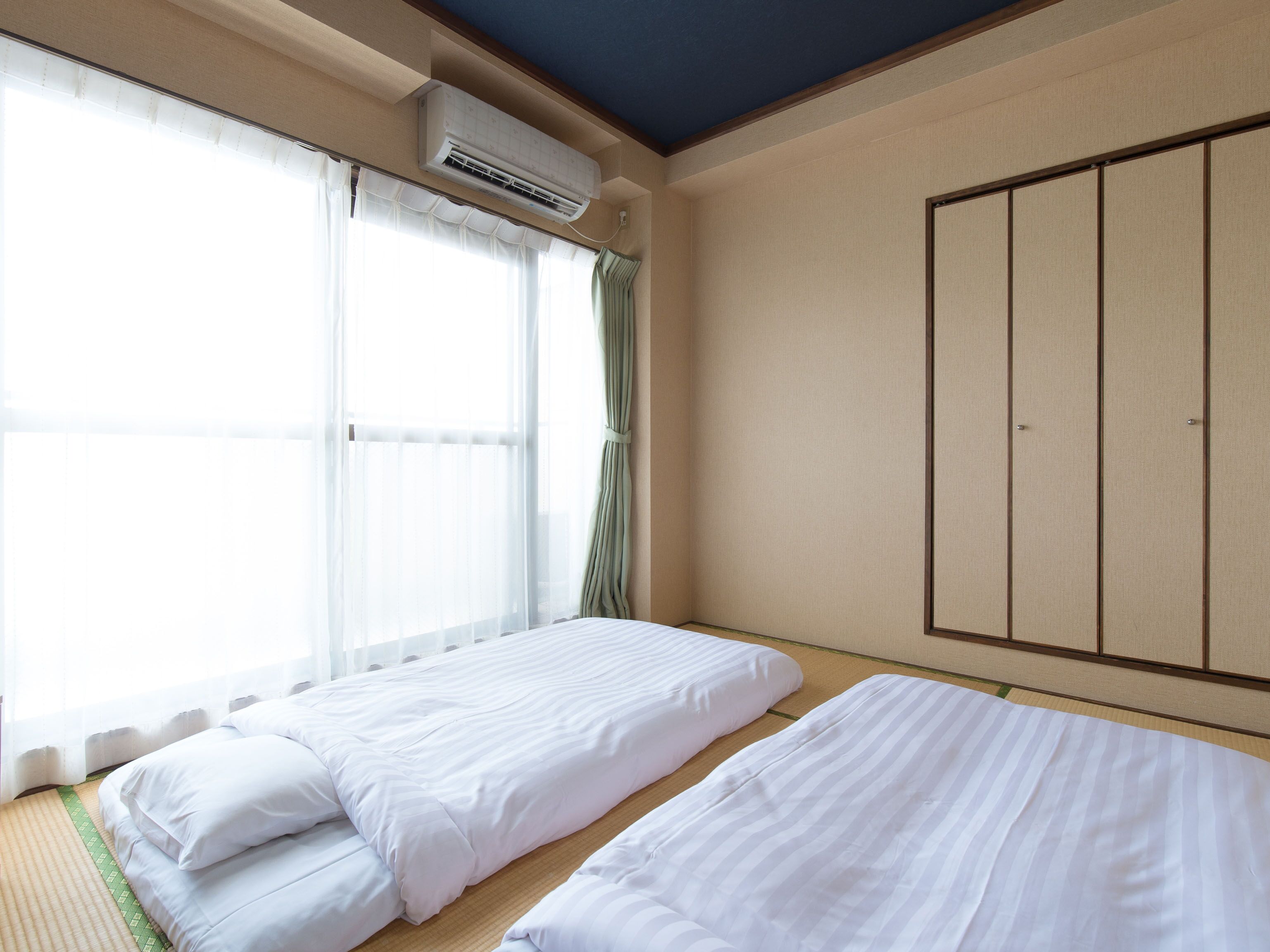 Una habitación con dos camas, un ventanal con cortinas translúcidas, un aire acondicionado y un armario.