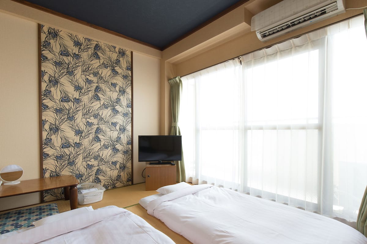 Habitació d'hotel amb dos llits, una televisió sobre un suport de fusta, una paret amb un disseny floral i una finestra gran amb cortines translúcides.