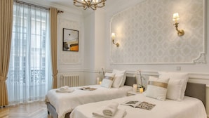 Ett hotellrum med två sängar, ett stort fönster med gardiner, ett element och väggmonterade lampor.