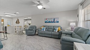 Ein modernes Wohnzimmer mit einem grauen Sofa, einer Essecke mit weißem Tisch und Stühlen sowie einer Küche im Hintergrund.