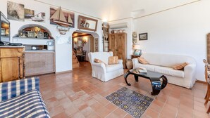 Un'ampia zona giorno con un divano bianco, un tavolino da caffè e un tappeto decorato. Si nota un mobiletto in legno e una scala che conduce in un'altra stanza.