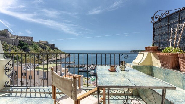 Un balcone con vista su una cittadina costiera, un tavolo con sedie e una pianta in vaso.