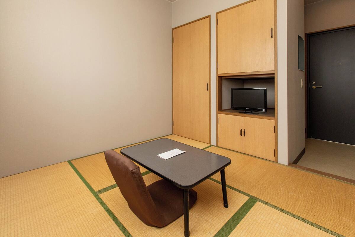 A small room with a tatami mat floor, a single chair, a table, a television on a shelf, and a door.