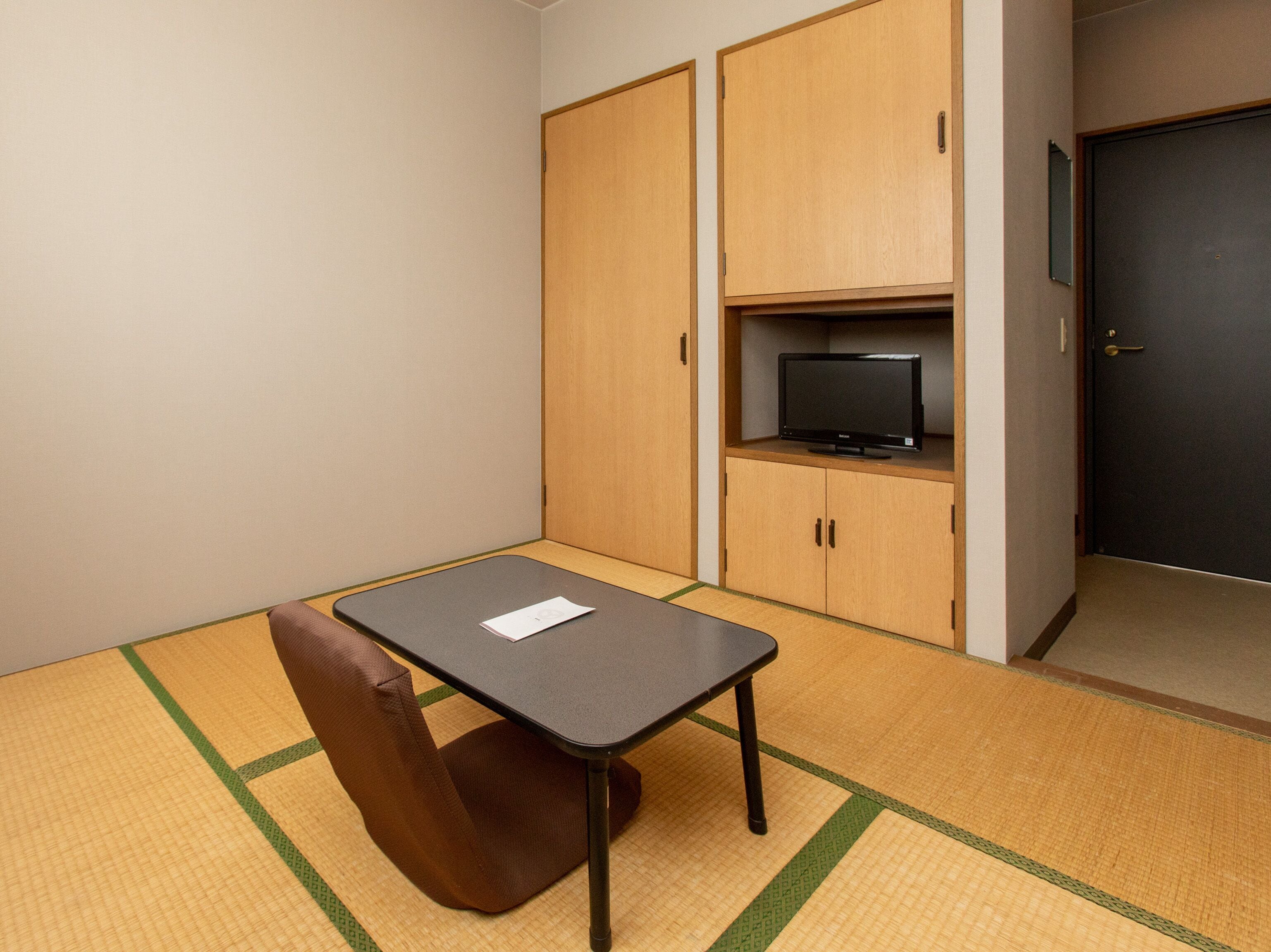 A small room with tatami mat flooring, a low table with a chair, wooden cabinets, and a television on a shelf.