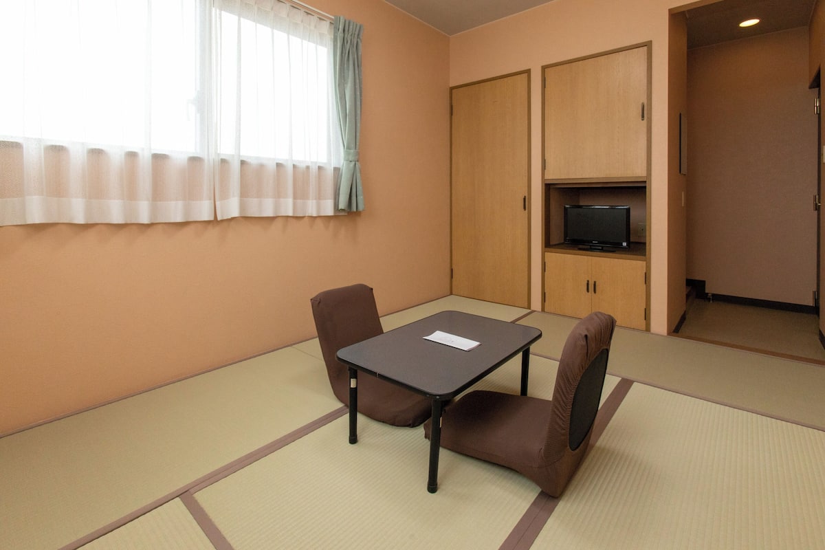 A room with tatami flooring, a table with chairs, a television on a shelf, and a door leading to another room.