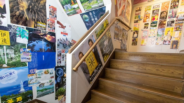 A wooden staircase with a handrail, surrounded by various posters and advertisements on the walls. - Tabist Business Ryokan Duck Ishinomaki Hebita (Isinomaki)