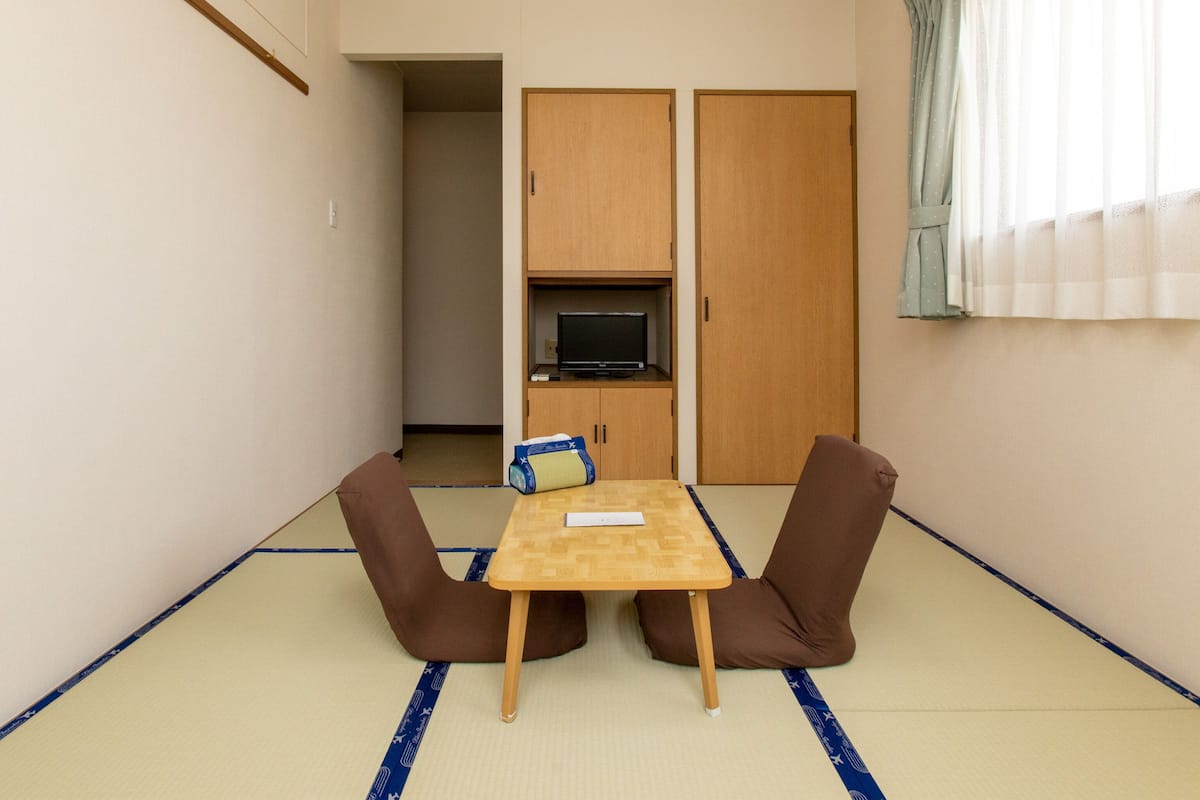 A small room with tatami flooring, a low table with two chairs, a television on a shelf, and sliding doors.