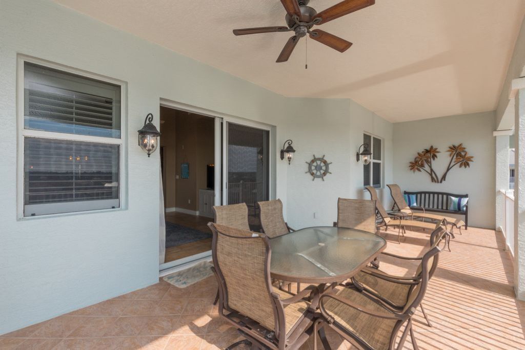 A covered patio with a glass table and wicker chairs, a ceiling fan, and a bench with decorative plants.