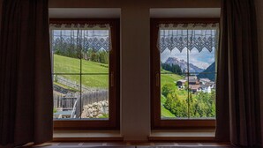 Duas janelas com cortinas de renda, cada uma com vista para uma paisagem verde com cerca de madeira e casas.