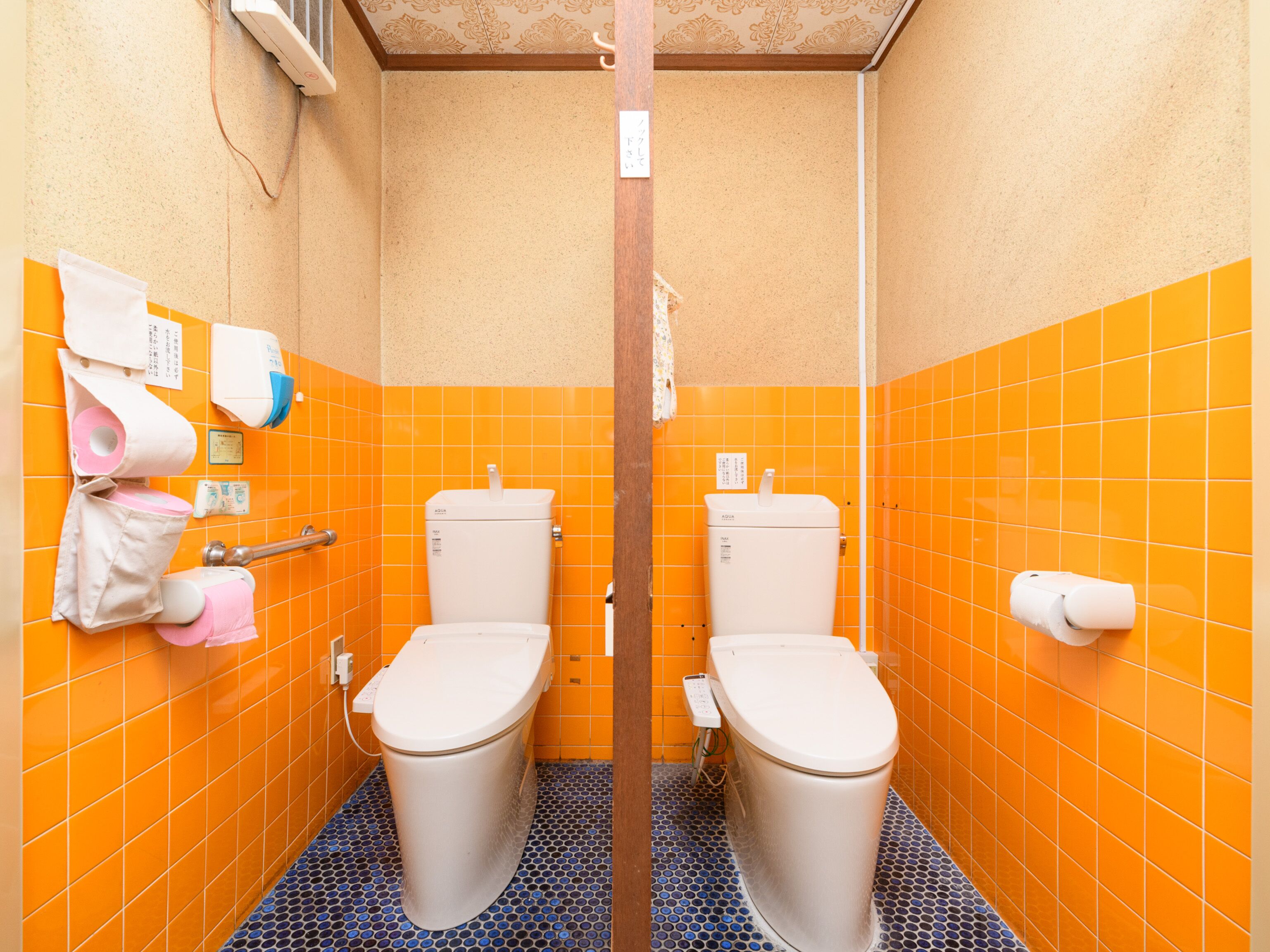 A bathroom with two toilets, orange tiled walls, and a blue patterned floor.