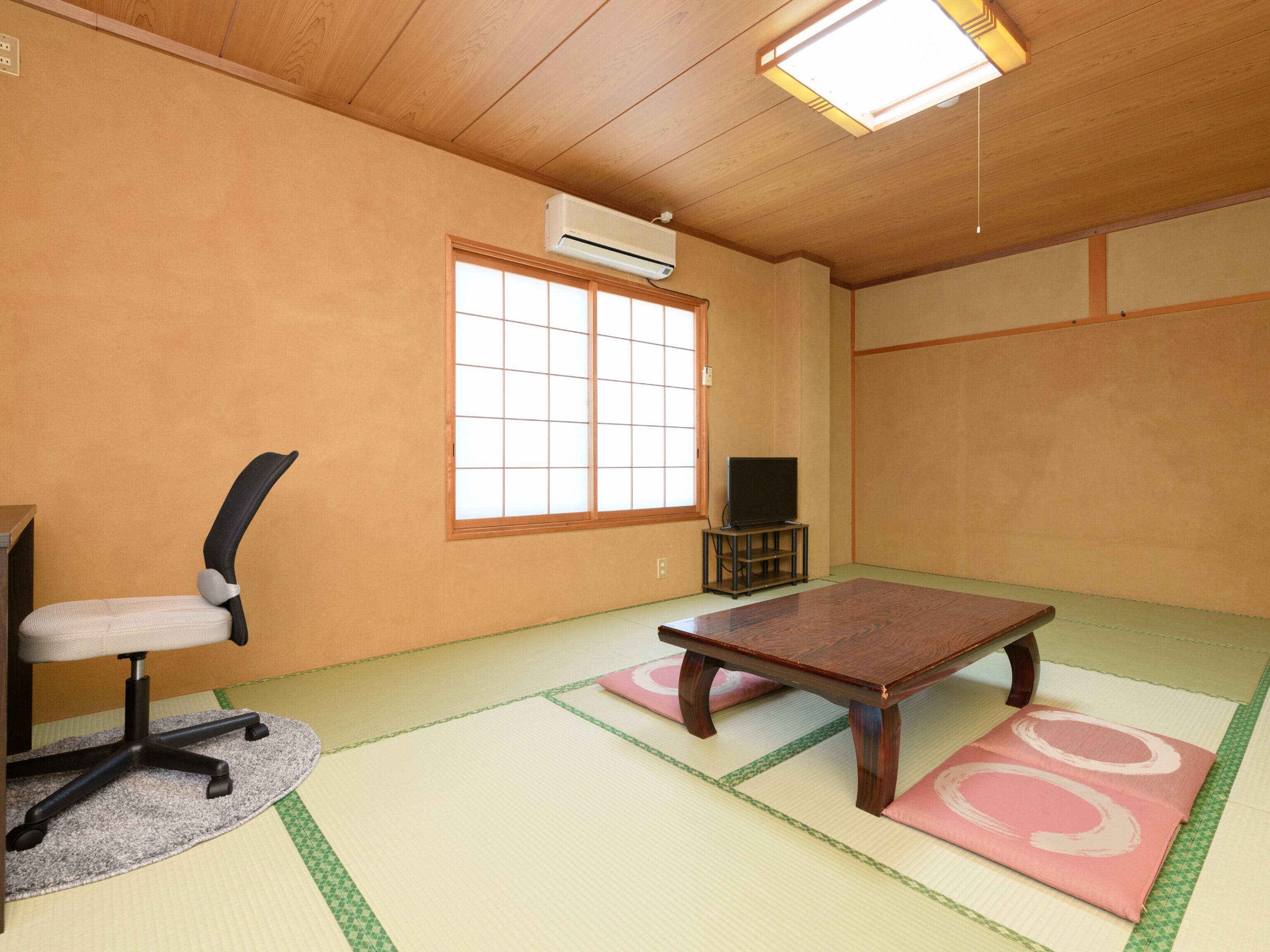 Una tradizionale stanza giapponese con pavimento in tatami, un tavolino in tatami, una sedia da ufficio nera, un televisore su un mobile e un condizionatore d'aria a parete.
