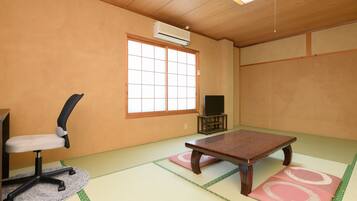 Una tradizionale stanza giapponese con pavimento in tatami, un tavolino in tatami, una sedia da ufficio nera, un televisore su un mobile e un condizionatore d'aria a parete.