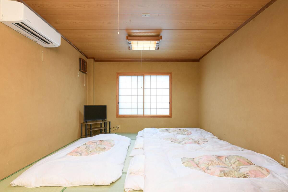 Une pièce avec un sol recouvert de tatamis, un futon avec une literie à motifs floraux, un téléviseur posé sur un petit meuble et une fenêtre avec des cadres en bois.