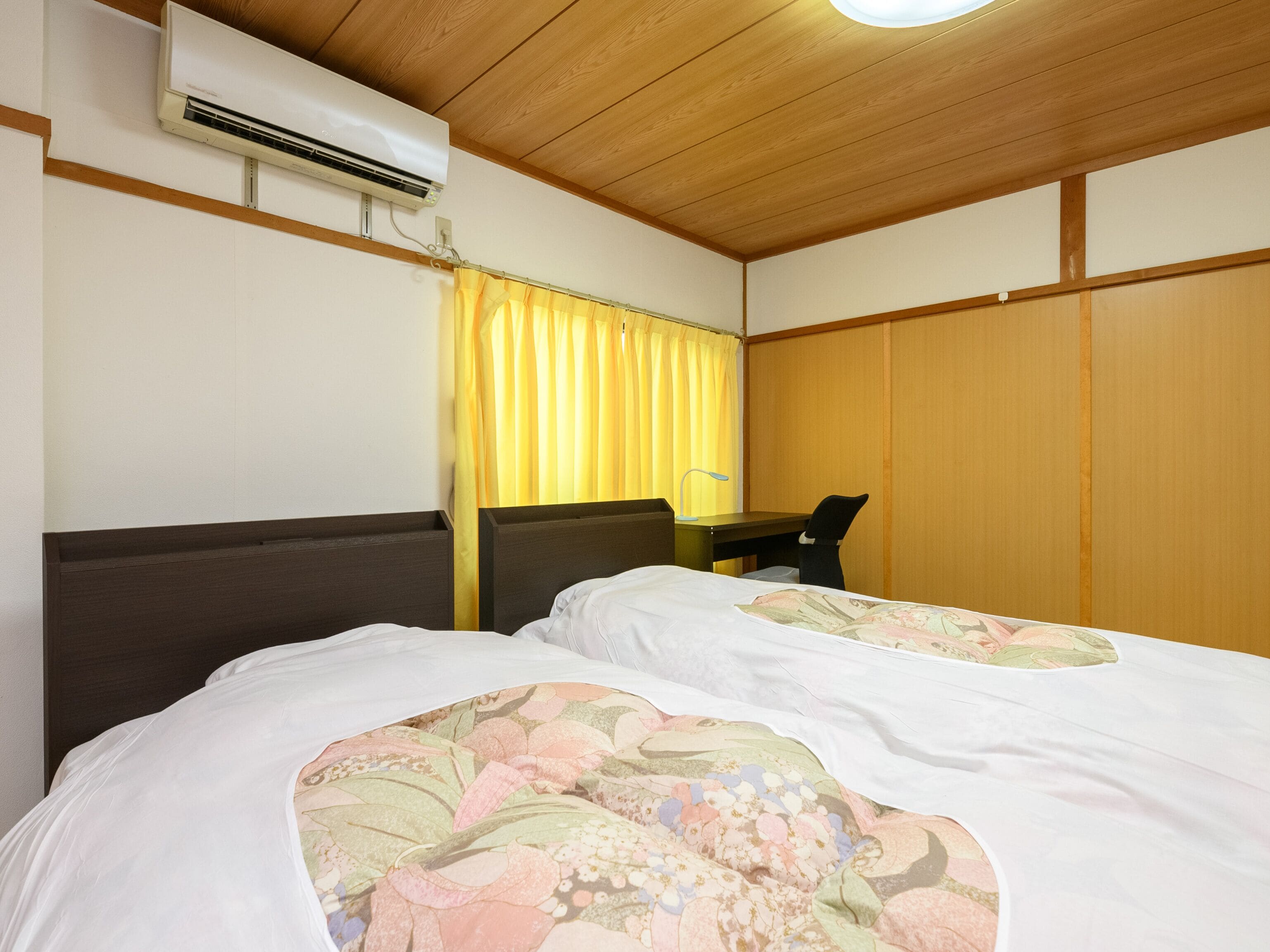 A hotel room with two beds, a desk, a chair, an air conditioning unit, and a curtain.