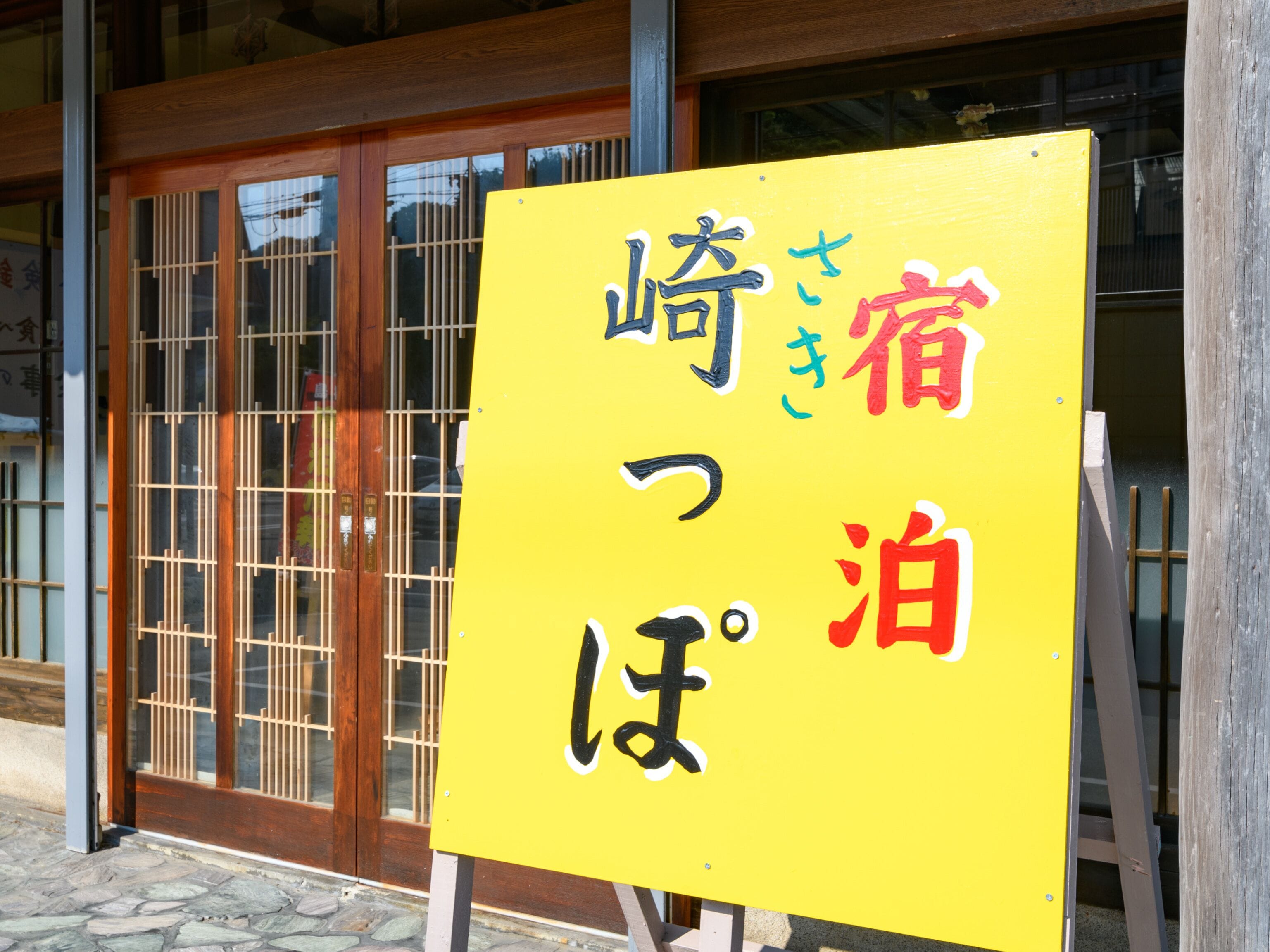 A yellow sign with Japanese characters, mounted on a metal frame, in front of a building with wooden doors and windows.