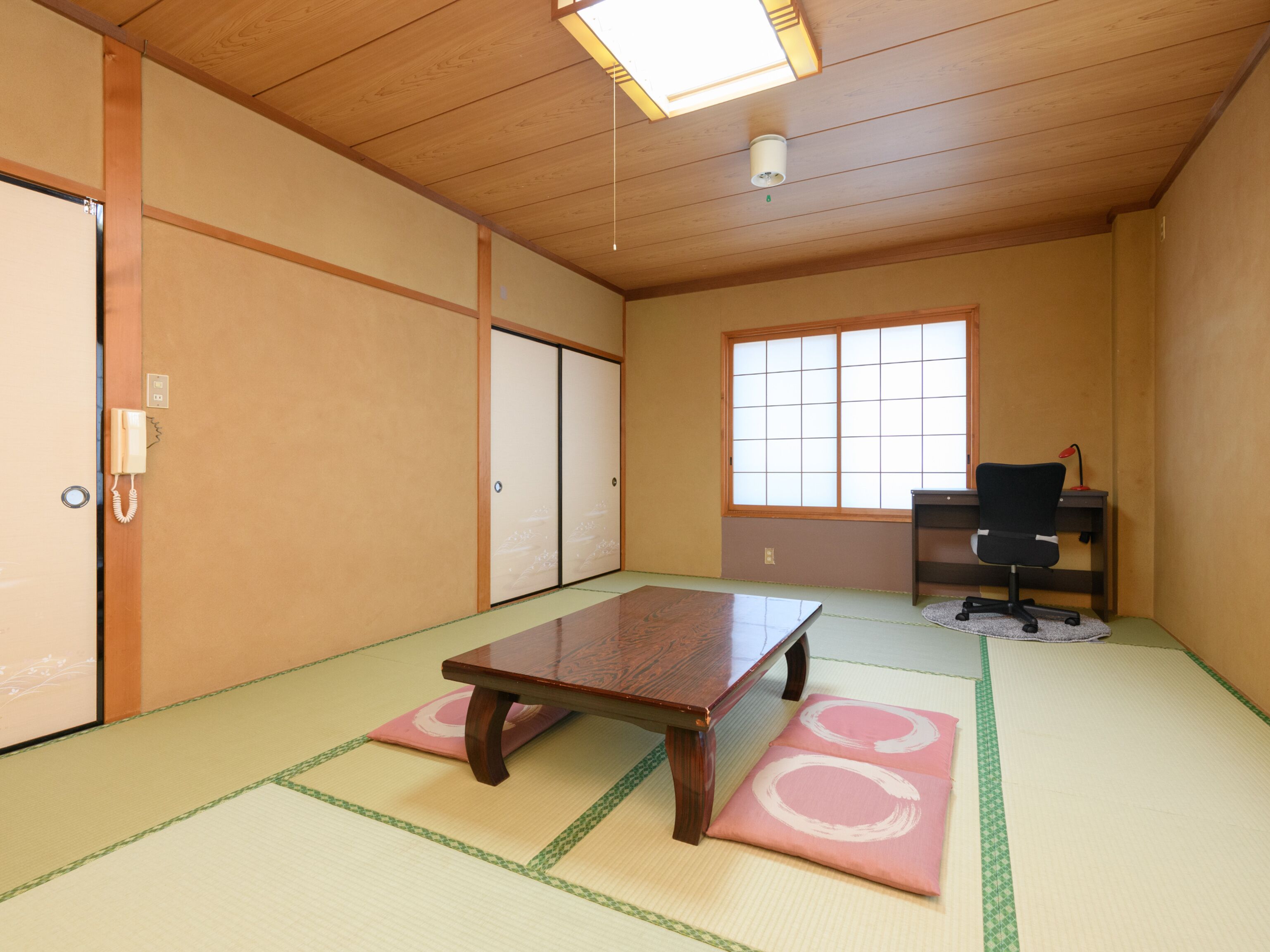 A traditional Japanese room with tatami flooring, a wooden table, sliding doors, a desk with a chair, and a window.