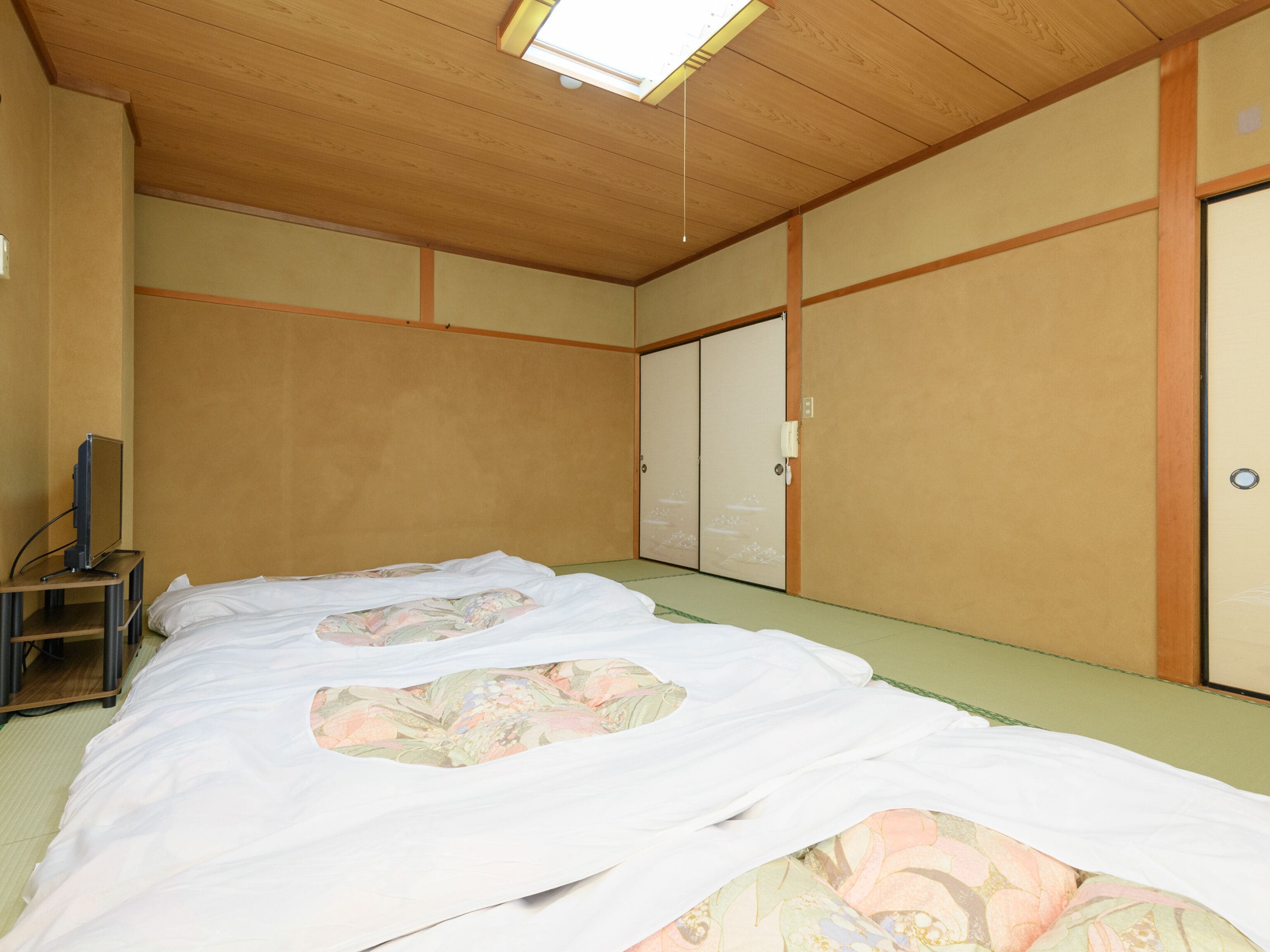 Een traditionele Japanse kamer met tatami vloerbedekking, een bed met bloemenprint beddengoed, een televisie op een standaard en schuifdeuren.