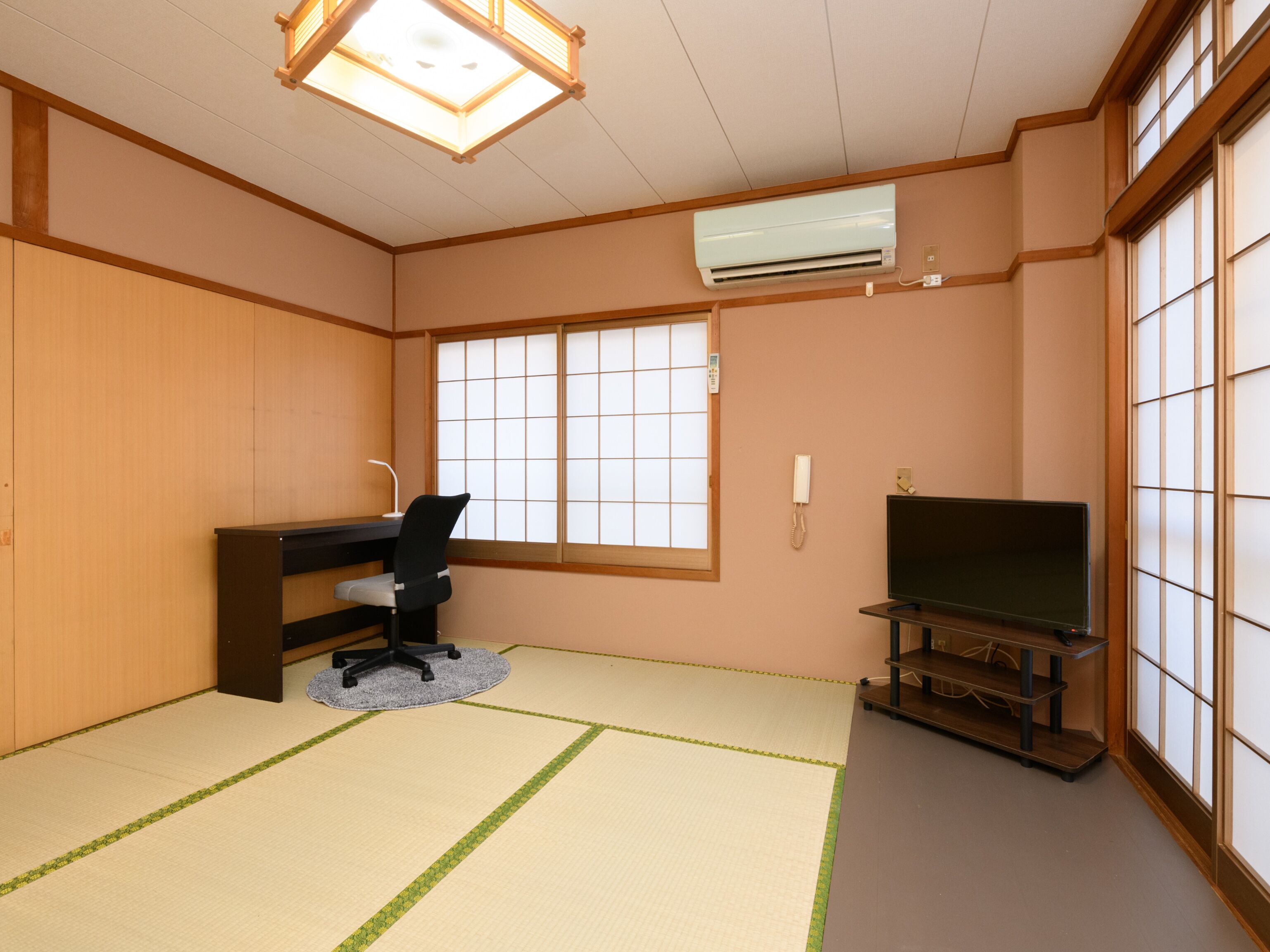 A traditional Japanese room with tatami flooring, a desk with a chair, a television, and a sliding door.