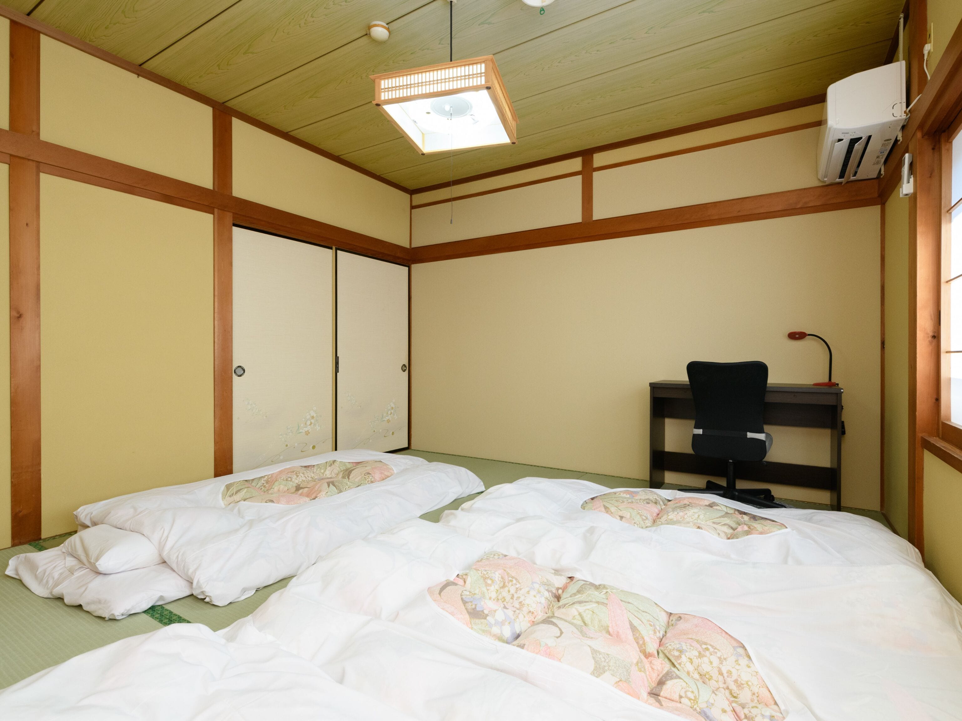 A room with two beds, a desk, and a chair. There is a ceiling fan and an air conditioning unit.