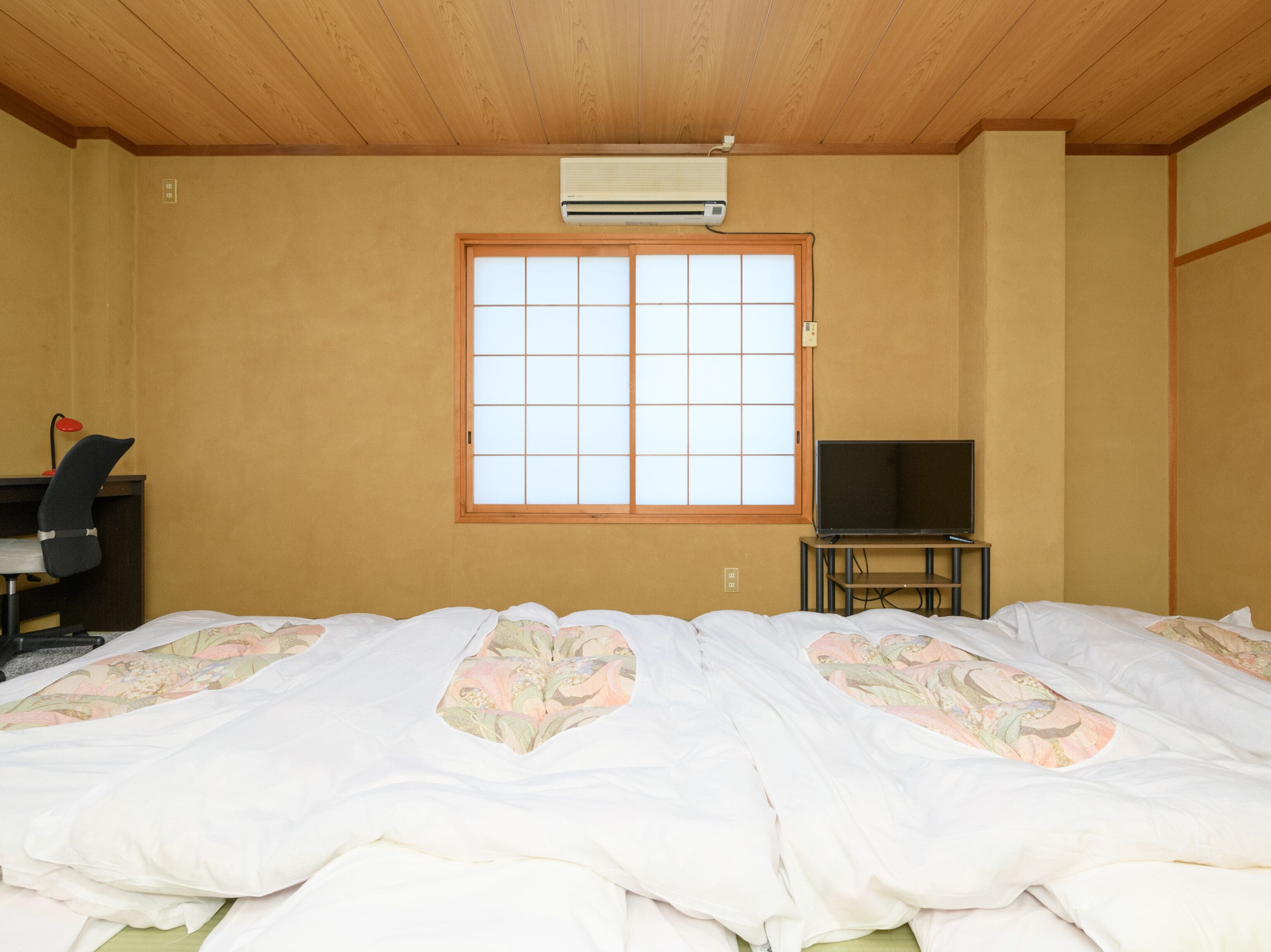 A hotel room with a wooden ceiling, a large window with a grid pattern, a bed with floral bedding, a desk with a chair, and a television on a stand.