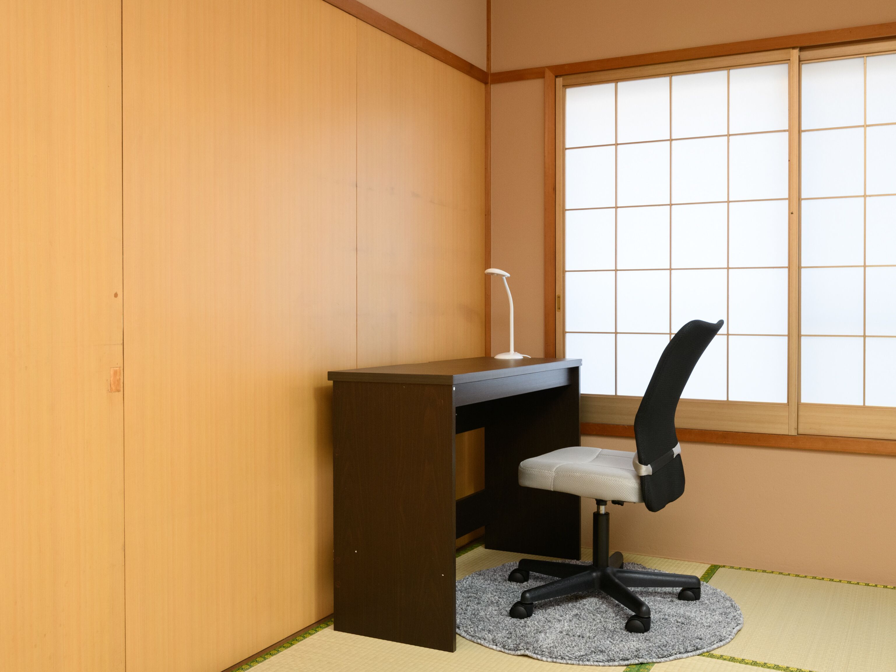 A small room with a wooden desk, a black office chair, a white desk lamp, and a large window with a grid pattern.
