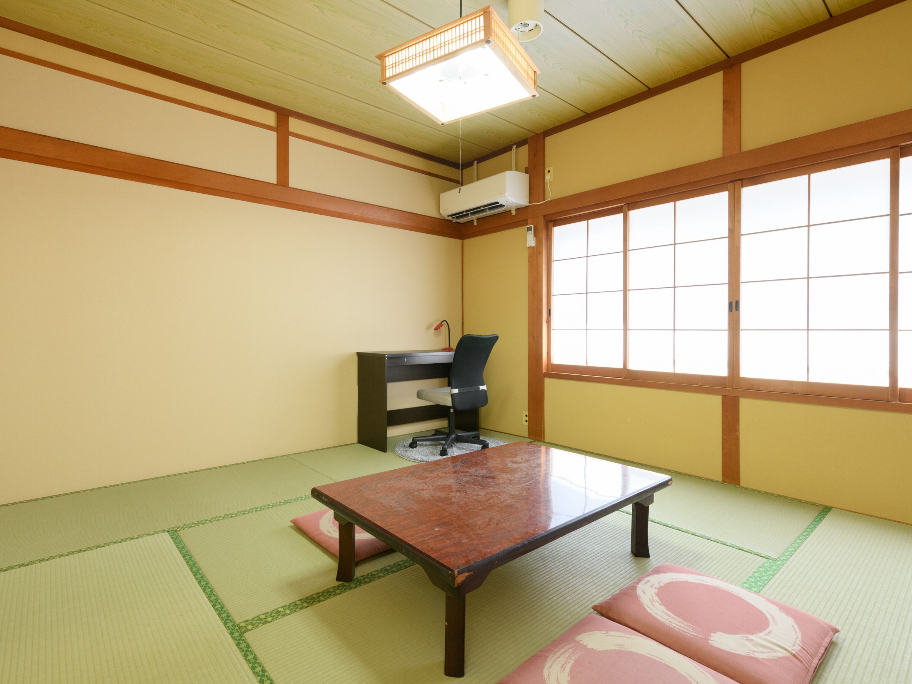 A traditional Japanese room with tatami flooring, a wooden table, a desk with a chair, and a large window.