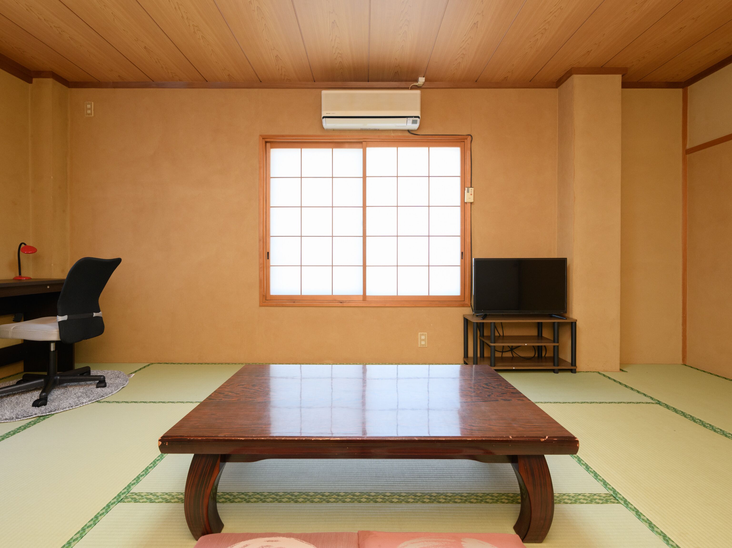 Une pièce traditionnelle avec tatamis, comprenant une table en bois, des nattes de tatami, un téléviseur posé sur un meuble, un bureau avec une chaise et une fenêtre à claustra de style shoji.