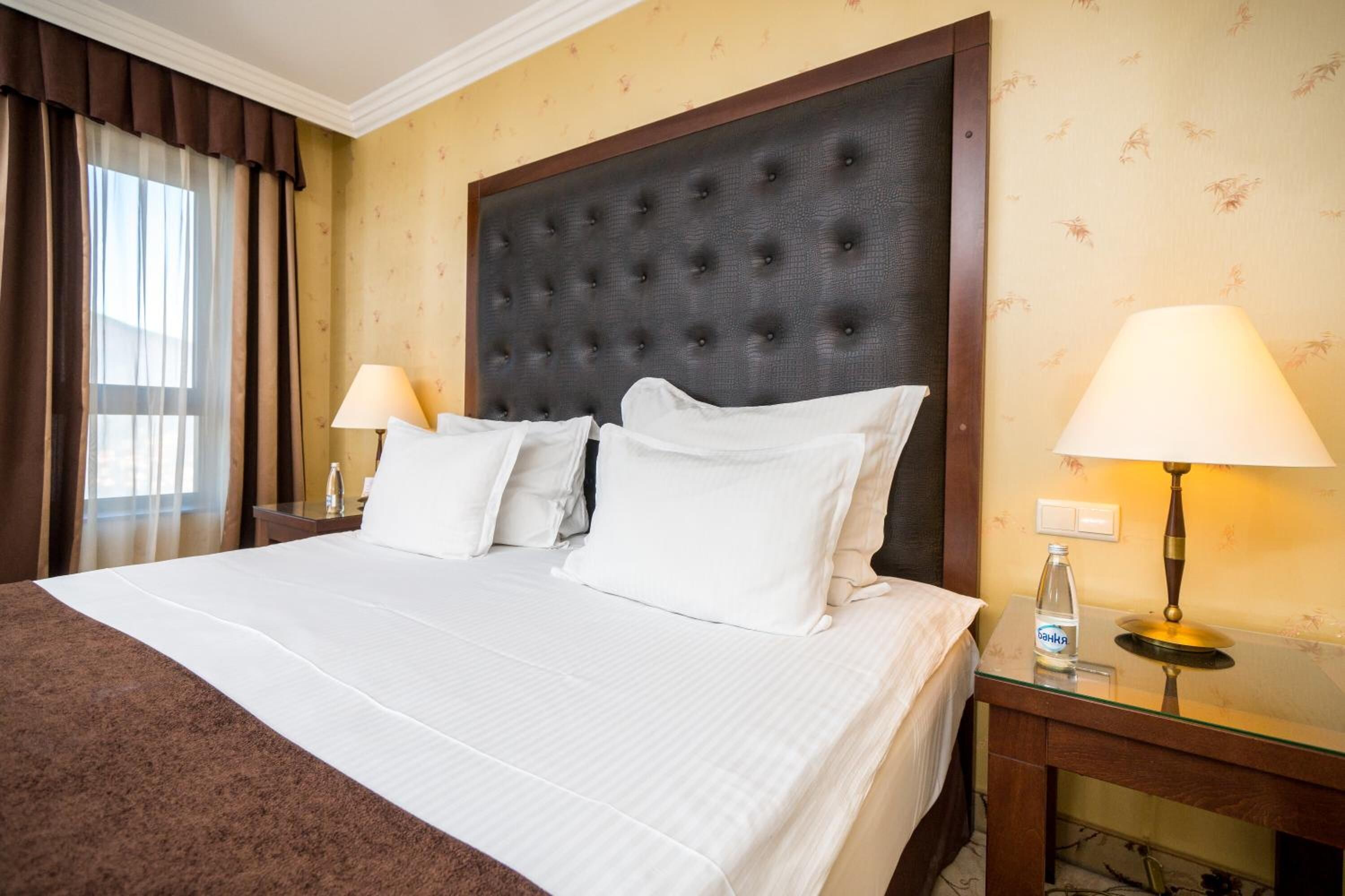 A hotel room with a large bed, two bedside tables, a headboard, and a window with curtains.