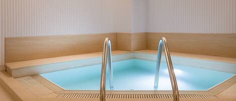 Jacuzzi de esquina con base de vidrio transparente, rodeado de paredes color beige y un banco de madera.