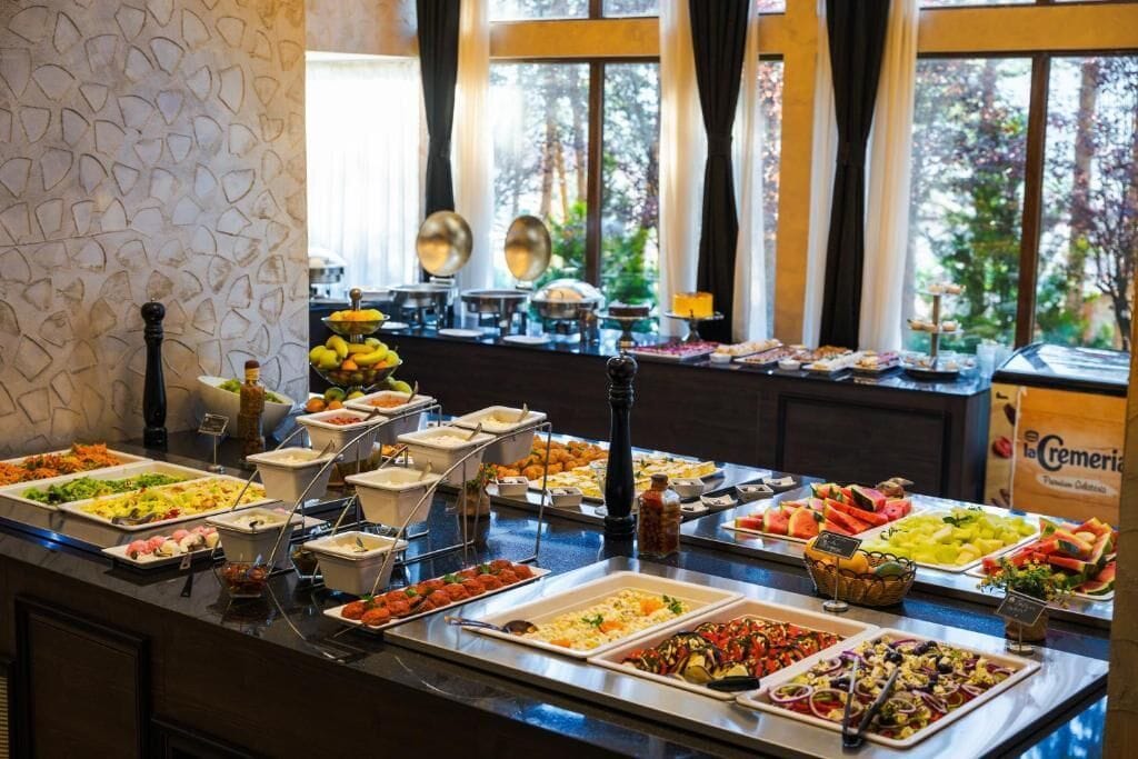 A buffet with various food options, including fruits, vegetables, and prepared dishes, set against a backdrop of large windows with a view of trees.