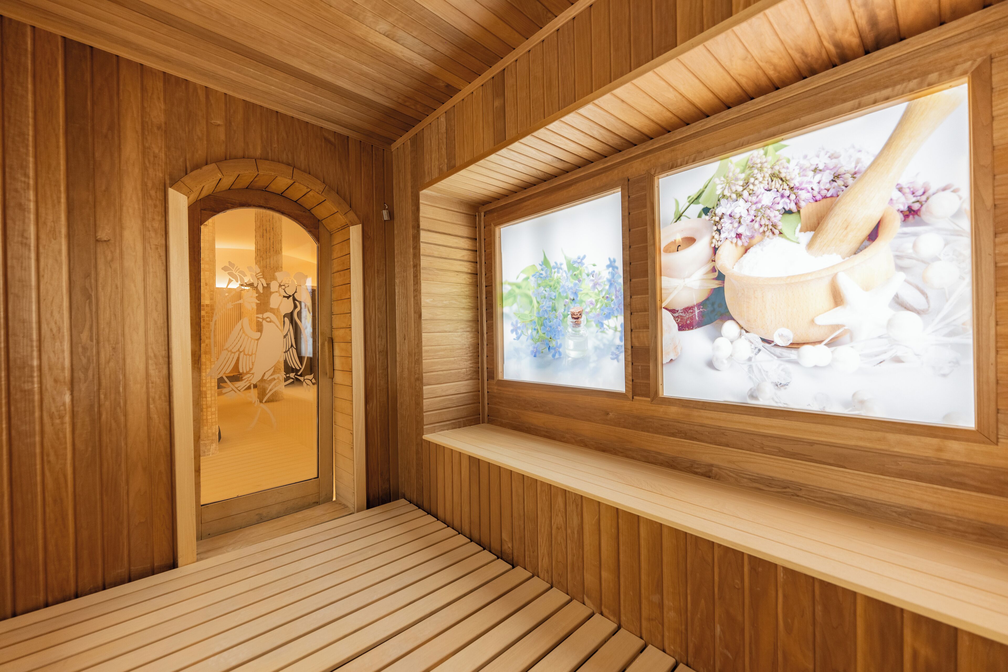 A wooden spa room with a bench, a wooden door, and a window with a view of flowers and a wooden mortar.