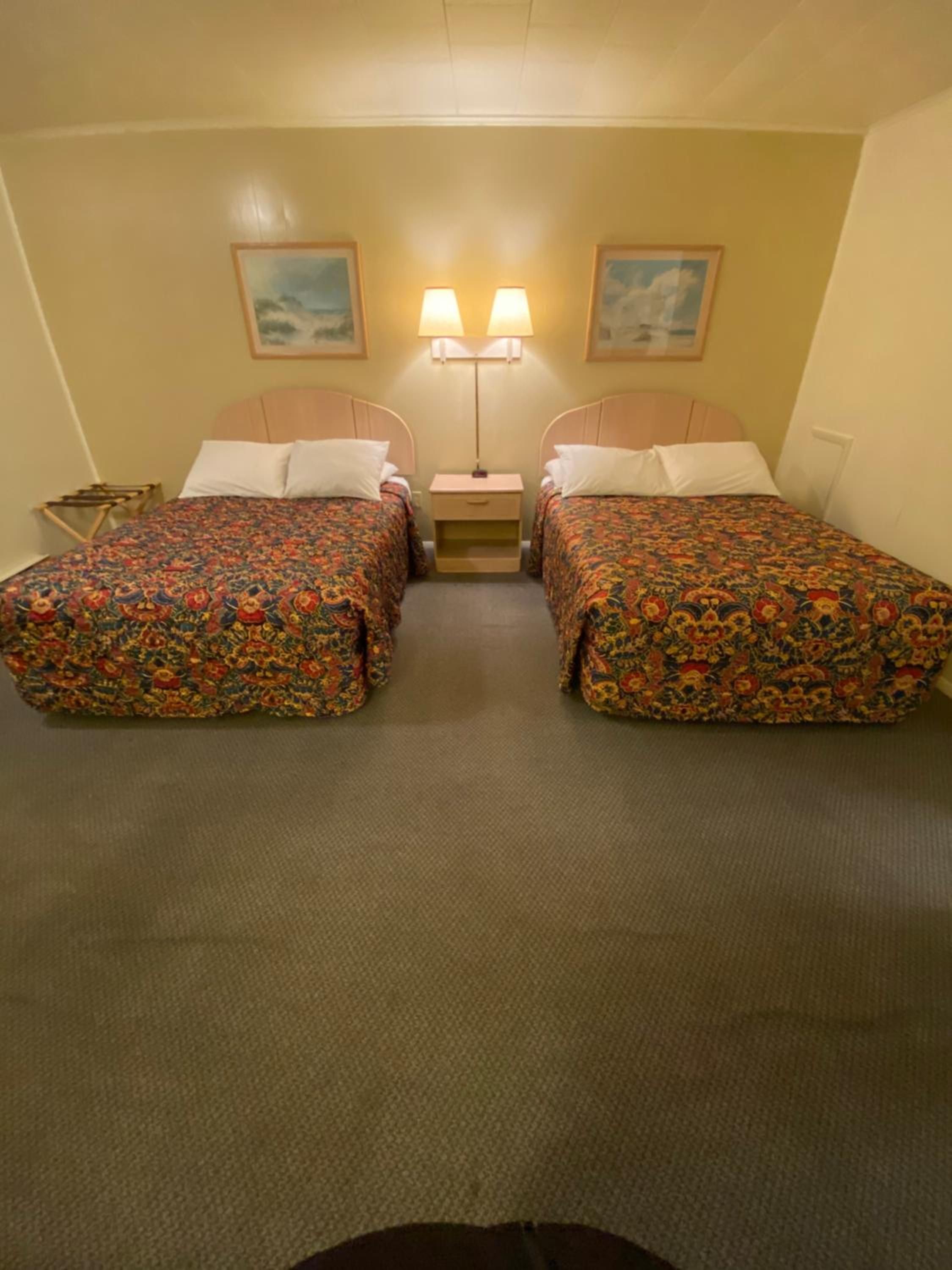 Habitación de hotel con dos camas, una mesita de noche, dos lámparas de pared y dos cuadros en las paredes.