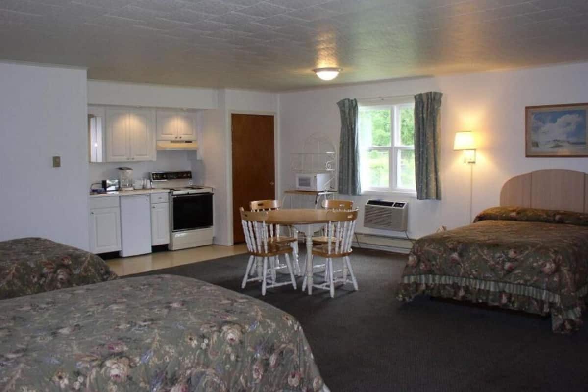 A room with two beds, a kitchenette, a dining table with chairs, a window with curtains, and a lamp.