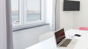 A room with a white table, a laptop, and a smartphone on it. There is a window with curtains and a view of the sea. - Fisherman's house in the harbor of Procida (Procida)