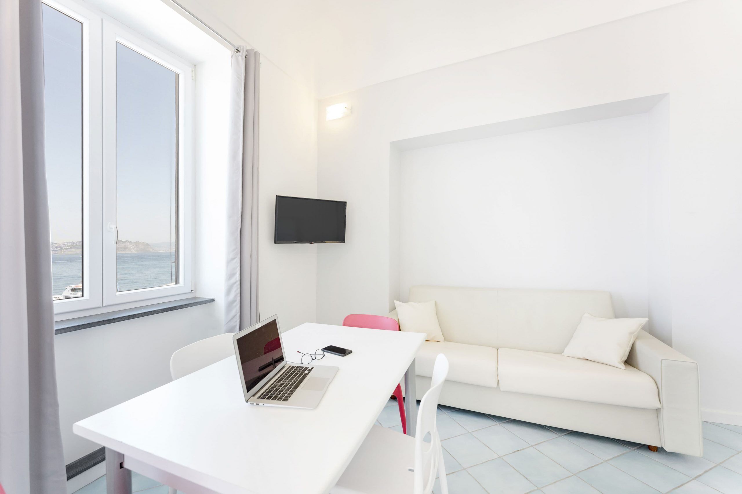 A modern room with a white desk, a laptop, a smartphone, a white sofa, and a TV mounted on the wall.